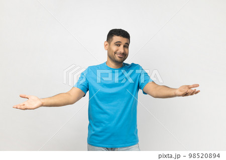 Portrait of unshaven man wearing blue T- shirt standing shows welcome gesture, spreads hands as wants to cuddle best friend, smiles broadly. Indoor studio shot isolated on gray background. Portrait of unshaven man wearing blue T- shirt standing shows welcome gesture, spreads hands as wants to cuddle best friend, smiles broadly. Indoor studio shot isolated on gray background. 98520894