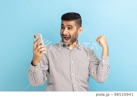 Portrait of bearded businessman holding cell phone and clenched fist, betting and winning, using mobile network services, wearing striped shirt. Indoor studio shot isolated on blue background. Portrait of bearded businessman holding cell phone and clenched fist, betting and winning, using mobile network services, wearing striped shirt. Indoor studio shot isolated on blue background. 98520941