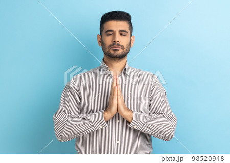 Portrait of calm relaxed young businessman with beard standing doing yoga meditating exercise, keeping palms together, wearing striped shirt. Indoor studio shot isolated on blue background. 98520948