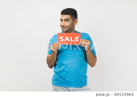 Portrait of positive unshaven man wearing blue T- shirt standing holding sale card in hands looking at camera with toothy smile, black friday. Indoor studio shot isolated on gray background. 98520969