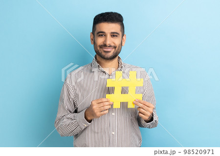 Viral hashtag and successful blogging. Satisfied businessman with beard holding large yellow hash symbol, wearing striped shirt. Indoor studio shot isolated on blue background. Viral hashtag and successful blogging. Satisfied businessman with beard holding large yellow hash symbol, wearing striped shirt. Indoor studio shot isolated on blue background. 98520971