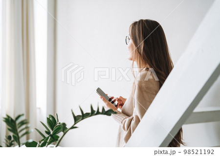 Smiling woman with long hair in beige cardigan drinking coffe and using mobile phone at home Smiling woman with long hair in beige cardigan drinking coffe and using mobile phone at home 98521192