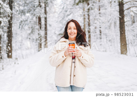 Smiling woman with paper cup of coffee in hands against background of snowy winter forest 98523031