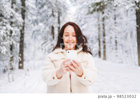 Smiling woman in winter clothes using mobile phone against background of snowy winter forest 98523032