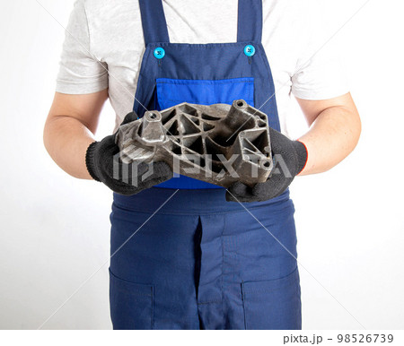 A car mechanic in blue overalls holds a pillow of a car engine in his hands. The concept of replacing an aluminum engine mount in a car, an automotive spare part. Close-up 98526739
