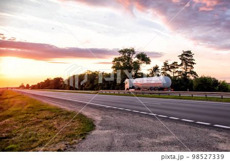 A truck with a semi-trailer tanker transports dangerous goods against the backdrop of a sunny sunset and the sky in the clouds. Transportation of fluid and liquid cargo in trucking industry 98527339