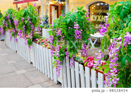 Outdoor cafe on street decorated artificial  purple flowers Ivy wisteria, green plant, white fence. Cafe summer terrace. Text translation: author's cuisine. 98528454
