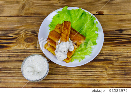 Fried fish fingers on a plate with lettuce and tartar sauce on wooden table. Top view 98528973