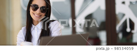 Smiling sun woman holding coffee and working on laptop in cafe Smiling sun woman holding coffee and working on laptop in cafe 98534059