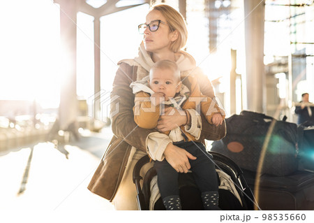 Motherat travelling with his infant baby boy child, walking, pushing baby stroller and luggage cart in front of airport terminal station Motherat travelling with his infant baby boy child, walking, pushing baby stroller and luggage cart in front of airport terminal station 98535660