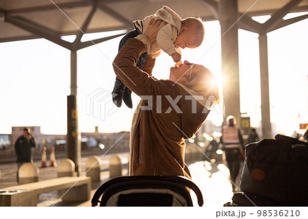 Motherat happily holding and lifting his infant baby boy child in the air after being rejunited in front of airport terminal station. Baby travel concept 98536210