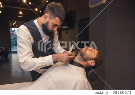 Professional barber carefully trimming the clients beard Professional barber carefully trimming the clients beard 98536671