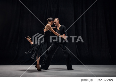 Man and woman, professional tango dancers performing in black stage costumes over black background. 98537269