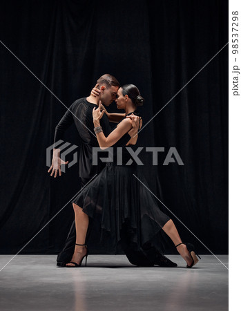 Man and woman, professional tango dancers performing in black stage costumes over black background. Attraction of movements 98537298