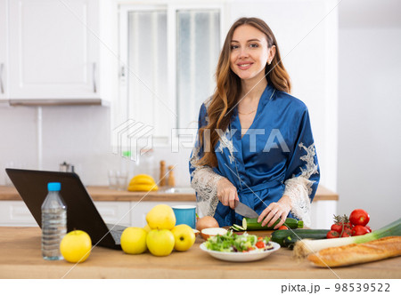 Young woman in blue robe cooking and watching TV series on laptop Young woman in blue robe cooking and watching TV series on laptop 98539522