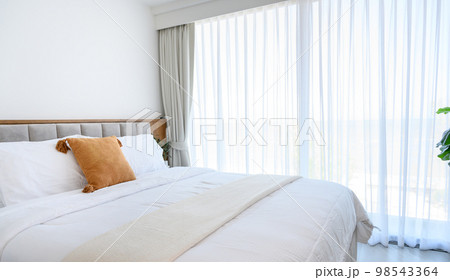 Modern interior cozy bed near a window with white curtains in bedroom. pillows and white soft blanket for rest and sleep. 98543364
