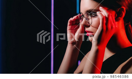 Retro optic. 90s look. 2000s eyewear fashion. Red purple color neon light closeup portrait of teen girl model face wearing round glasses on dark black copy space background. 98546386
