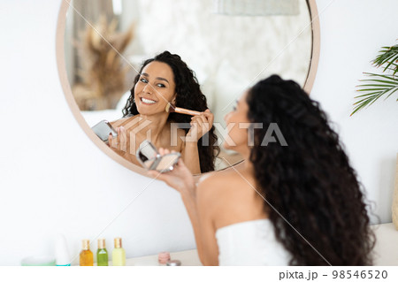 Smiling pretty arab millennial curly lady in towel applying blush on face, looking in mirror, enjoy daily procedure Smiling pretty arab millennial curly lady in towel applying blush on face, looking in mirror, enjoy daily procedure 98546520