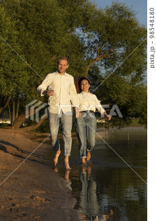 Caucasian man runs holding Latin woman hand on beach, happy couple enjoys time together, having fun near water. Green trees on background. Lovers wear casual clothes. Vertical plane 98548180