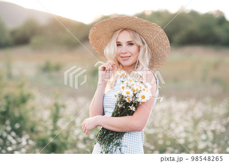 Smiling beautiful blonde woman 25-29 year old holding bouquet of fresh daisy wearing straw hat and dress posing in meadow close up. 98548265
