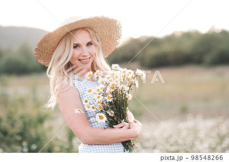 Smiling beautiful blonde woman 25-29 year old holding bouquet of fresh daisy wearing straw hat and dress posing in meadow close up. 98548266