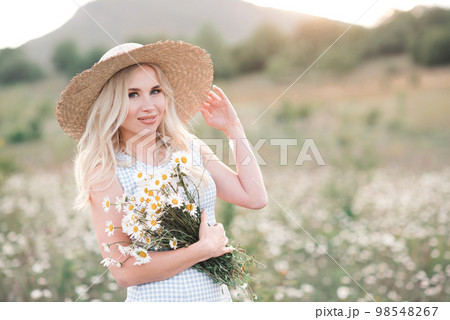Smiling beautiful blonde woman 25-29 year old holding bouquet of fresh daisy wearing straw hat and dress posing in meadow close up. Smiling beautiful blonde woman 25-29 year old holding bouquet of fresh daisy wearing straw hat and dress posing in meadow close up. 98548267