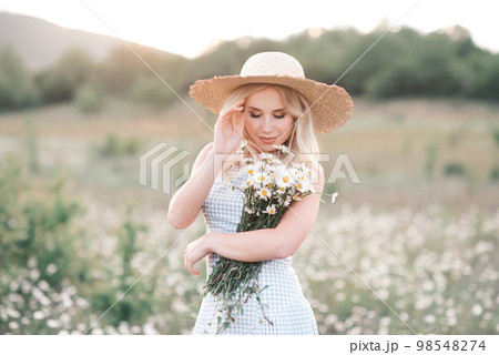 Smiling beautiful blonde woman 25-29 year old holding bouquet of fresh daisy wearing straw hat and dress posing in meadow close up. 98548274