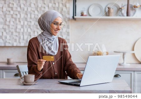 Cheerful and successful woman in hijab at home in kitchen with laptop and bank credit card makes online purchase in online store, Muslim woman chooses products and gifts remotely. 98549044