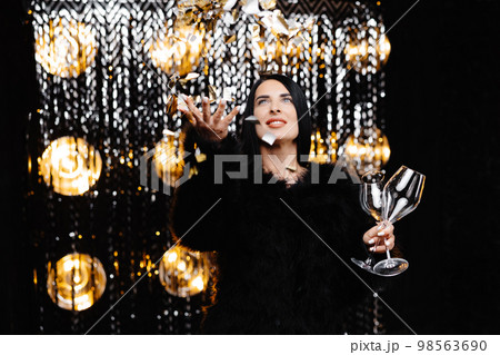Happy woman with brunette long hair in trendy evening fur coat holding glass and confetti on shine wall in studio Happy woman with brunette long hair in trendy evening fur coat holding glass and confetti on shine wall in studio 98563690