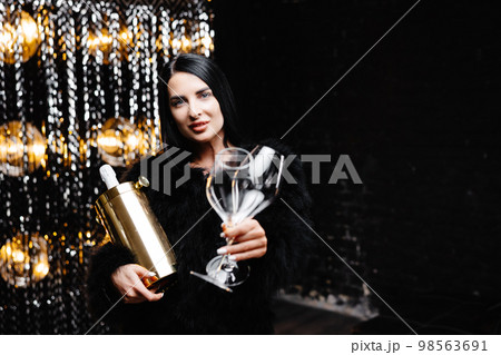 Happy woman with brunette long hair in trendy evening fur coat holding glass and bucket with bottle of champagne on shine wall in studio Happy woman with brunette long hair in trendy evening fur coat holding glass and bucket with bottle of champagne on shine wall in studio 98563691