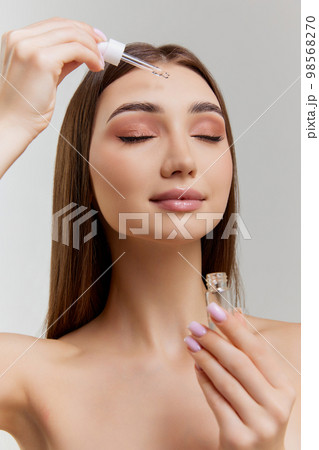 Organic products. Beautiful young girl applying face serum, taking care after skin over grey studio background. Concept of skincare and natural beauty Organic products. Beautiful young girl applying face serum, taking care after skin over grey studio background. Concept of skincare and natural beauty 98568270