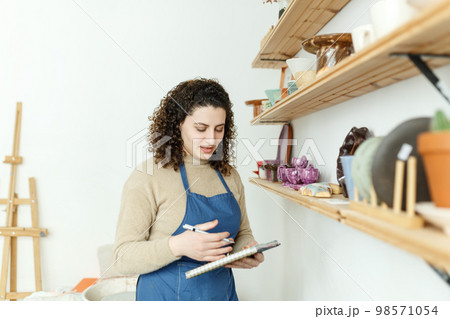 Happy young ceramist looking away thoughtfully while taking stock. Creative businesswoman managing a store with handmade ceramic products. Female entrepreneur running a successful small business 98571054