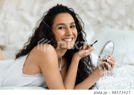 Cheerful arab millennial curly lady in domestic clothes lies on bed, doing basic makeup with mascara Cheerful arab millennial curly lady in domestic clothes lies on bed, doing basic makeup with mascara 98573427