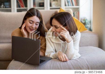 Happy pretty european millennial ladies in sweaters lie on sofa, watch video on laptop, surfing in internet Happy pretty european millennial ladies in sweaters lie on sofa, watch video on laptop, surfing in internet 98573466