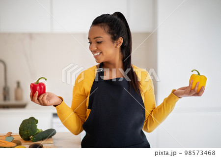 Cheerful millennial african american female in apron chooses peppers in hands at table with vegetables Cheerful millennial african american female in apron chooses peppers in hands at table with vegetables 98573468
