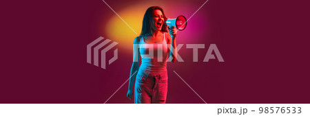 Emotive young girl in casual clothes shouting in megaphone over gradient dark pink background in neon light. Information, news. Banner, flyer Emotive young girl in casual clothes shouting in megaphone over gradient dark pink background in neon light. Information, news. Banner, flyer 98576533