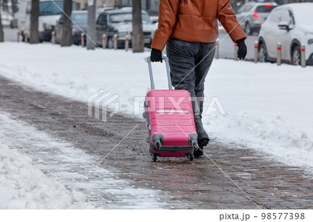 A traveler woman walks along a paved sidewalk in the winter season, carrying a travel bag on wheels. Copy space. 98577398