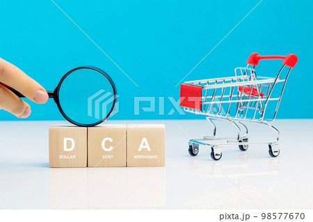 Magnifying glass in woman's hand and DCA word on a wooden cube in idea Dollar Cost Averaging investment strategy near empty shopping cart. Saving stock or savings on a monthly, quarterly basis. 98577670