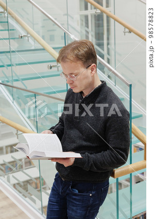 man in library is reading book sitting on steps of glass staircase man in library is reading book sitting on steps of glass staircase 98578823