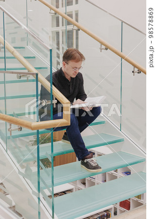 man in library is reading book sitting on steps of glass staircase man in library is reading book sitting on steps of glass staircase 98578869