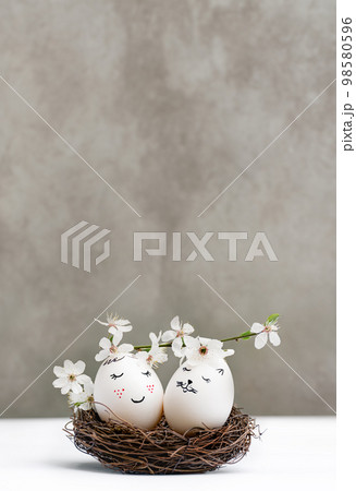 Happy Easter. White Easter eggs in wicker wooden nest on white table on gray background. View is straight. Blossoming branch of cherry with white flowers. Stylish spring template with copy space Happy Easter. White Easter eggs in wicker wooden nest on white table on gray background. View is straight. Blossoming branch of cherry with white flowers. Stylish spring template with copy space 98580596
