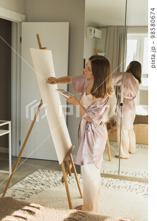 Young woman artist painting on canvas on the easel at home in bedroom - art and creativity concept Young woman artist painting on canvas on the easel at home in bedroom - art and creativity concept 98580674