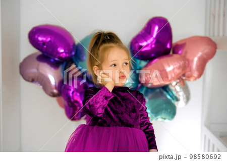 A thoughtful girl with pouting lips looks away, pressing her hand to her cheek. A child in a room with festive decorations made of heart-shaped balls on the wall 98580692