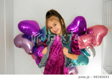 A stylish girl in a dress and a denim vest smiles sweetly, standing at a decorated wall with multicolored foil balloons in the shape of a heart. Decor in the children's room for Valentine's Day 98580693