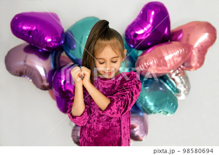 A girl in a purple velvet dress folded her arms in the shape of a heart, isolated against a background with bright multicolored balloons. Decor for Valentine's Day A girl in a purple velvet dress folded her arms in the shape of a heart, isolated against a background with bright multicolored balloons. Decor for Valentine's Day 98580694