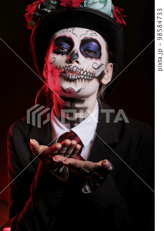 Woman doing kissy face with santa muerte make up, blowing air kisses at camera. Model wearing dios de los muertos make up and costume to celebrate mexican halloween holiday in studio. Woman doing kissy face with santa muerte make up, blowing air kisses at camera. Model wearing dios de los muertos make up and costume to celebrate mexican halloween holiday in studio. 98584733