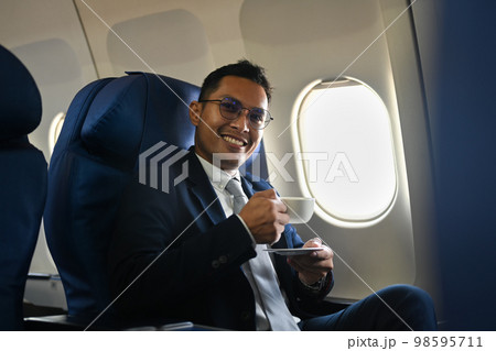 Positive businessman passenger drinking coffee from paper cup while sitting comfortable seat in airplane cabin Positive businessman passenger drinking coffee from paper cup while sitting comfortable seat in airplane cabin 98595711