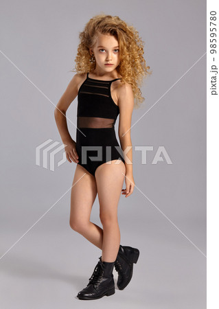 Beautiful little girl gymnast in a black sports swimsuit and boots on a gray background. Beautiful little girl gymnast in a black sports swimsuit and boots on a gray background. 98595780