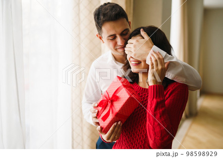 A young man gives his beloved a present and closes her eyes with a hand. Valentine's Day. 98598929