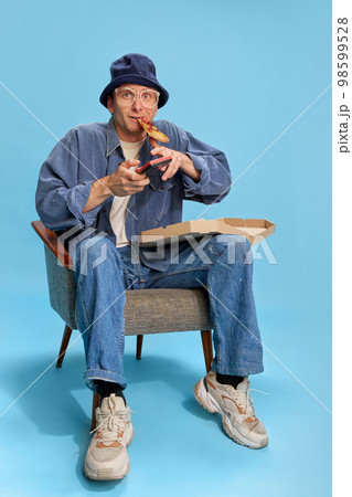 Into game. Cheerful man in jeans clothes sitting with pizza and playing retro game console over blue studio background. Concept of emotions, lifestyle, hobby 98599528